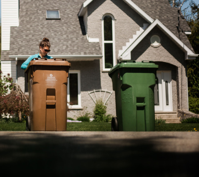 Femme debout près de son bac de composte et son bac de recyclage devant sa propriété